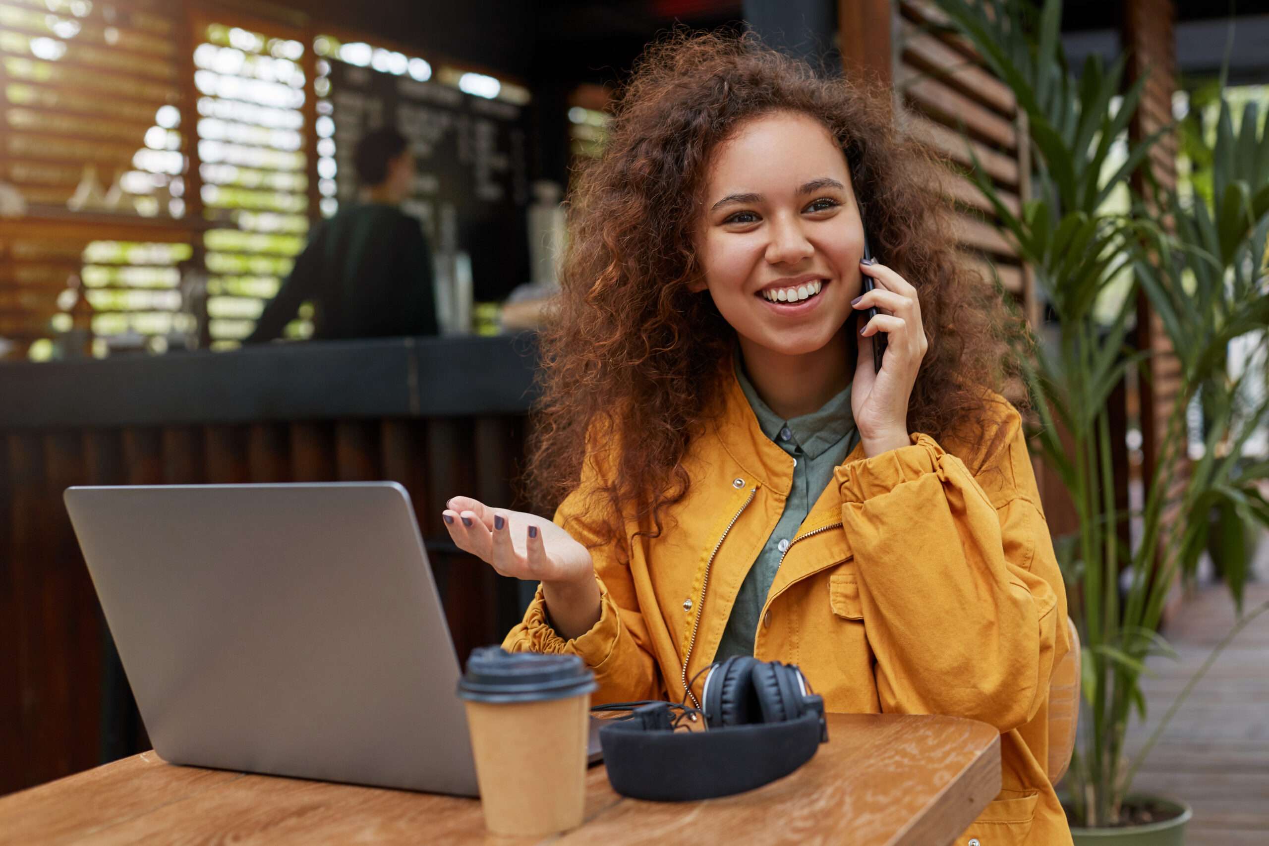 Photo Of Young Dark Skinned Curly Woman Siting On A Cafe Terrace Wearing In Yellow Coat